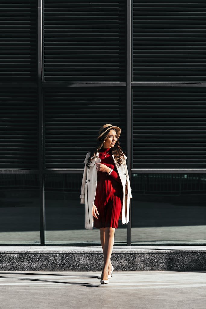 Full body trendy female with long curly hair in red dress and hat and high heeled shoes and coat adjusting collar while walking along road from contemporary building in daylight