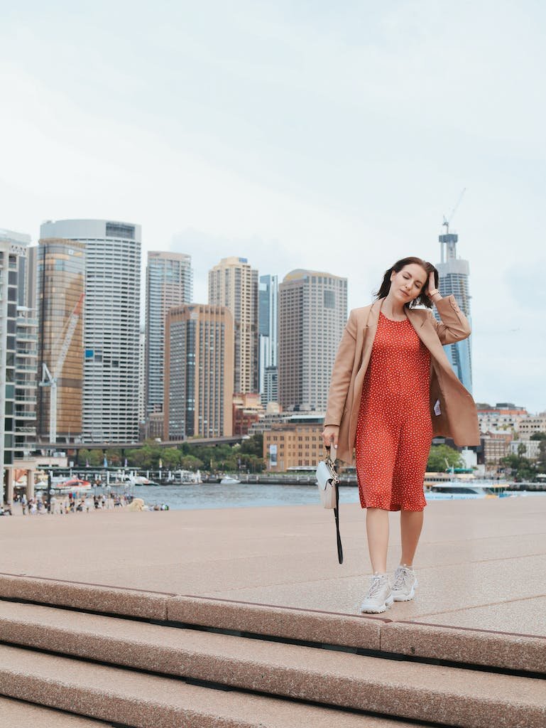 Full length of stylish young dreamy female in red dress and beige jacket strolling along embankment and touching hair against modern skyscrapers