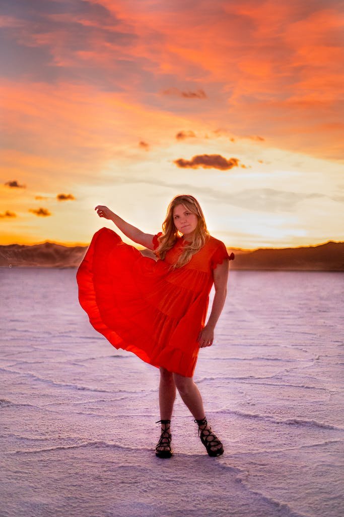Young female in bright flying dress standing with raised arm in desert under cloudy sky at sunset while looking at camera